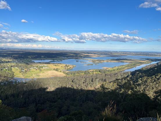 Hawkesbury Lookout, Australia Looking towards Sydney. Panoramic landscape featuring a broad view of a valley with a large, reflective lake in the center. The foreground includes grassy areas with rocks and shrubs. Dense forest covers the midground, leading to open fields and patches of greenery surrounding the lake. A horizon of distant rolling hills under a clear blue sky with scattered clouds.