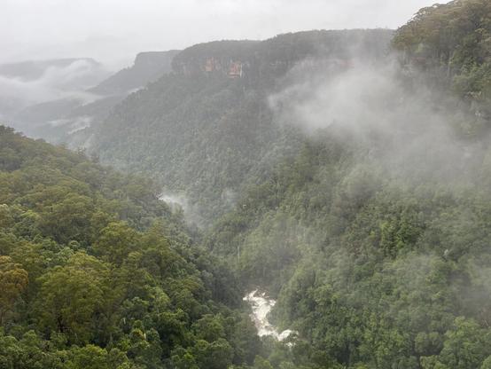 Yarrunga Valley, Australia Dense green forested mountains shrouded in mist, with a narrow river cutting through the valley. Fog drifts across the landscape, partially obscuring the view, while distant cliffs are visible through the haze.