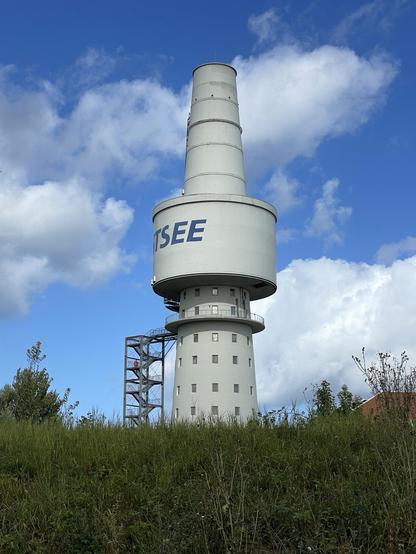 Ein großer, zylindrischer Turm steht unter einem blauen Himmel mit vereinzelten Wolken. Der Turm ist weiß. Eine Metalltreppe führt hinauf zu einer Plattform. Im Vordergrund sind saftiges grünes Gras und einige Bäume zu sehen.