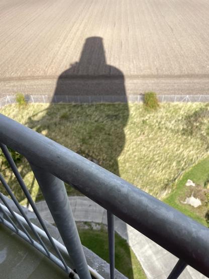 Ein Bild, das den Schatten des Aussichtsturms auf dem Boden zeigt, mit Blick auf ein Balkongeländer im Vordergrund und bewirtschafteten Feldern im Hintergrund.