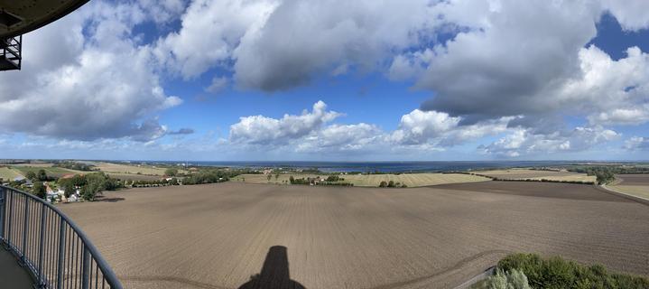 Ein Panoramablick auf offene Felder und Ackerland, die unter einem teilweise bewölkten blauen Himmel auf ein Gewässer treffen. Die Landschaft ist geprägt von einer Mischung aus Ackerland und Baumgruppen, wobei entlang des Wassers ein ferner Horizont sichtbar ist.