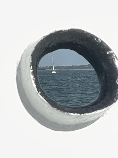 Ein Blick auf ein Segelboot auf dem Wasser durch eine runde Öffnung mit einer verschwommenen Küstenlinie im Hintergrund. Die Szene fängt eine friedliche maritime Atmosphäre ein.