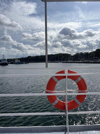 Ein roter Rettungsring hängt an der Reling eines Bootes mit Blick auf ruhiges Wasser und einen Yachthafen im Hintergrund. Der Himmel ist teilweise bewölkt, ein Hauch von Blau schimmert durch.