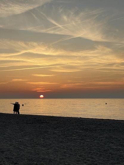 Silhouetten stehen bei Sonnenuntergang an einem Strand und blicken zum Horizont, wo die strahlende Sonne über dem ruhigen Wasser untergeht. Der Himmel ist in Orangetönen und sanften Wolken gehalten und schafft eine heitere Abendstimmung.