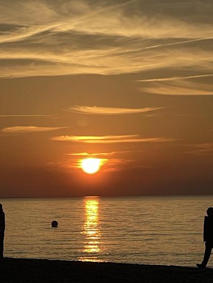 Ein atemberaubender Sonnenuntergang über ruhigem Wasser. Die Sonne steht tief am Himmel und spiegelt sich auf der Wasseroberfläche. Am Strand sind die Silhouetten zweier Menschen zu sehen. Der Himmel ist mit weichen Wolken bedeckt, die in warmen Orangetönen gemalt sind.