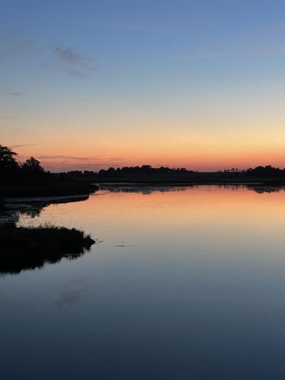 Eine ruhige Landschaft mit einem ruhigen Gewässer, in dem sich ein farbenfroher Sonnenuntergang spiegelt. Der Himmel wechselt von Blau- zu Orange- und Rosatönen, und entlang des Wasserrands sind Silhouetten von Bäumen und Land zu erkennen.