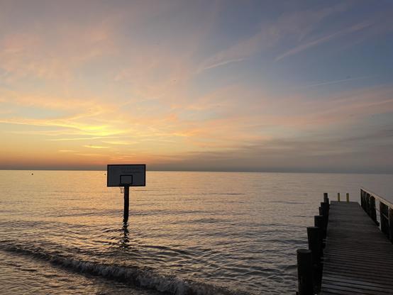 Eine ruhige Uferszene bei Sonnenuntergang. Das ruhige Wasser spiegelt den farbenfrohen Himmel wider. Im Vordergrund steht ein Basketballkorb auf einer Stange in der Nähe des Wassers. Ein Holzsteg ragt in den See hinein, einige Pfosten sind sichtbar.