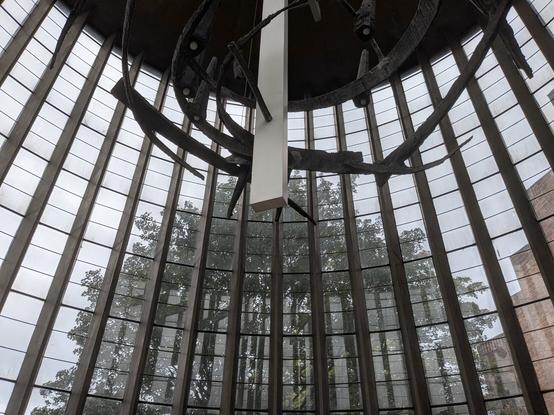 Photo of a large, tall curved glass window divided by thin vertical concrete pillars with a tree seen outside plus some castle-like brick buttresses on the LHS. Overhead is a large, angular black metal sculpture in a spiral shape. The colours are muted with an overcast sky so the whole image appears almost monochrome.