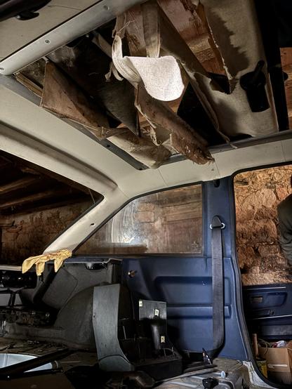 An interior view of a vehicle showing a damaged ceiling with torn fabric and wood, along with some disassembled parts on the dashboard and a visible window. The setting appears old and unkempt, with stone walls in the background.