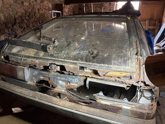 A rusted and deteriorating rear view of an old car, showing significant corrosion and damage, stored in a dimly lit area with a stone wall in the background. Dust covers the surface, and various items are scattered around, suggesting neglect.