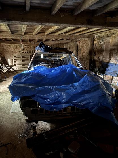 An unoccupied vehicle is covered with a blue tarp inside a rustic wooden structure. The surrounding area has various objects, including a chair and some tools, highlighting a cluttered garage or workshop environment. Natural light enters the space, illuminating parts of the interior