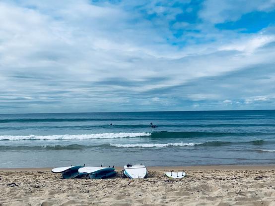 Manly Beach, Australia Surfboards resting on sandy beach with ocean waves in the background. Two surfers in the water appear near the horizon. Overhead, a mostly cloudy sky with patches of blue.