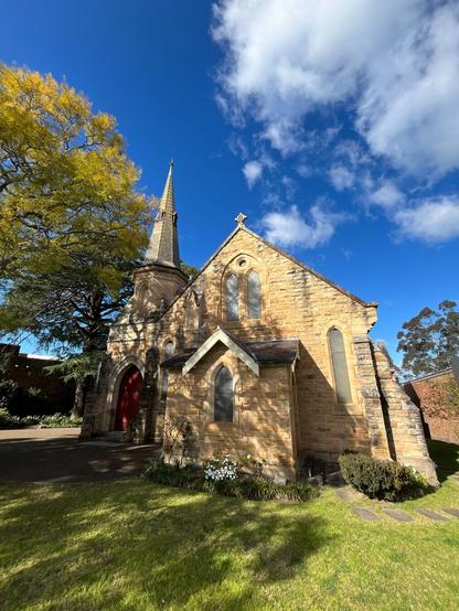 Springwood Presbyterian Church. Springwood Australia Stone church with tall, pointed steeple and red door, set against a bright blue sky with fluffy clouds. Yellowing tree on the left, casting shadows on the building. Foreground includes well-maintained grassy area and white flowers.