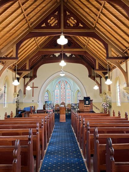 Wooden church interior featuring arched ceiling with exposed beams, aligned pews along a central aisle with a blue carpet, stained glass windows in the background, and a pulpit adorned with flowers on the right. Hanging lights illuminate the space, and a wooden lectern is positioned near the center.