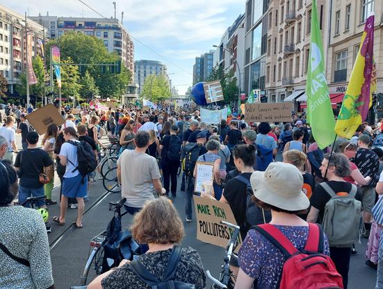 Die Friedrichstraße auf Höhe des Friedrichstadtpalastes bis zur Weidendammer Brücke voller Menschen mit Schildern, Fahnen und/oder Fahrrädern und/oder Rucksäcken. Alle warten, daß es weiter geht - viele unterhalten sich oder schauen sich um. Rechts und links die Fassaden verschiedener Epochen - mit Stuck oder Glasfronten oder Plattenbau-Elementen. Die bunte Mischung der Berliner Innenstadt. Weit im Hintergrund die S-Bahnbrücke über die Friedrichstraße.