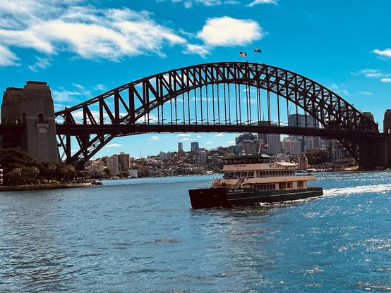 Sydney Harbour Bridge spans across the water under a clear blue sky. A ferry travels on the water in the foreground, with the city's skyline visible in the background.