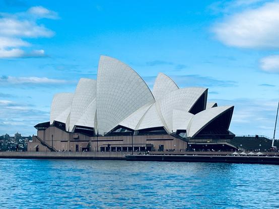 Sydney Opera House with its iconic white, sail-like roof structures set against a blue sky, located at the edge of a body of water. A few fluffy clouds are scattered across the sky. The building's unique architectural design is complemented by its reflection in the water. Spectators and visitors are visible around the structure's base.