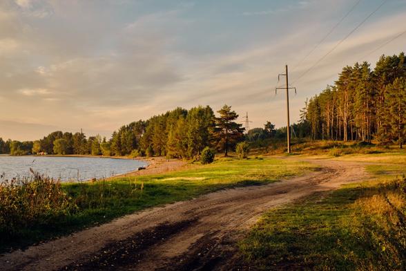 A peaceful lakeside scene featuring a sandy pathway winding through green grass and trees. Power lines stretch across the sky, and the calm water reflects the soft light of the rising sun.