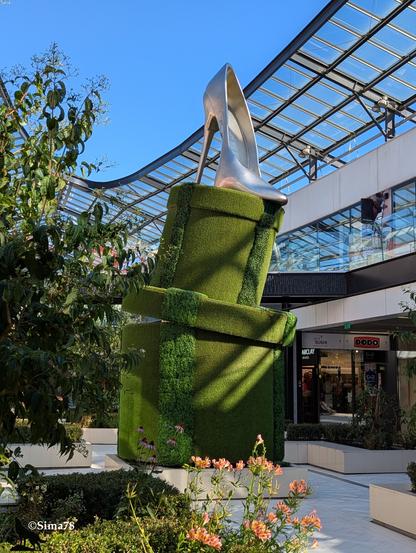 Sculpture végétale en forme de boite ouverte surmontée d’un escarpin géant en métal, exposée dans un centre commercial sous une verrière.
A plant sculpture in the shape of an open box topped with a giant metal stiletto, displayed in a shopping mall under a glass roof.
Escultura vegetal en forma de caja abierta rematada por un zapato de tacón gigante de metal, expuesta en un centro comercial bajo una claraboya.