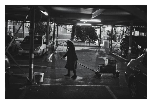 Black and white, low-angle photo of a grandma pulling a small cart with two boxes on it. She is walking from the right to the left side of the frame in an elevated parking garage. In the background, there are parked cars and a person walking. The parking garage is illuminated by fluorescent lights.