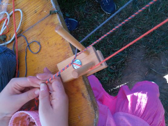 Kinderhände verdrillen Garne zu einem dünnen Seil. Man sieht eine hölzerne Hilfseinrichtung, die an einem Tisch klemmt.