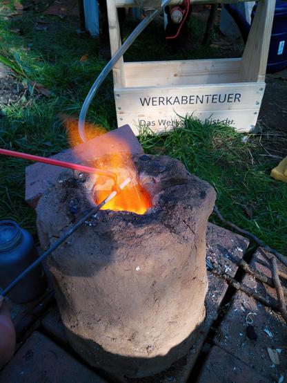 In der Stichflamme eines kleinen Lehmofens wird eine Glasperle auf einen Dorn gewickelt. Es wird eine rote Perle. Hinten steht eine Holzkiste mit dem Aufdruck "Werkabenteuer - Das Werkstück bist du".