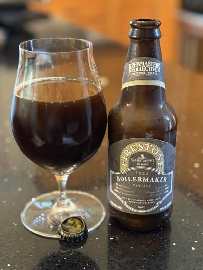 A tall snifter glass full of medium dark brown beer. The 12 ounce brown bottle has a black labels with white lettering and a gold arch with “Firestone” in white letters. The black bottle cap with a gold bear is down front