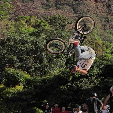 Fotografia de um homem jovem, com uma calça jeans, camiseta regata, capacete, um tênis vermelho, fazendo uma manobra em uma bicicleta de BMX, em que ele está no ar de cabeça para baixo. Ao fundo muitas árvores e algumas pessoas o assistindo.