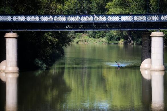 se ve un puente sobre un ancho río, y entre sus dos columnas está pasando una piragua