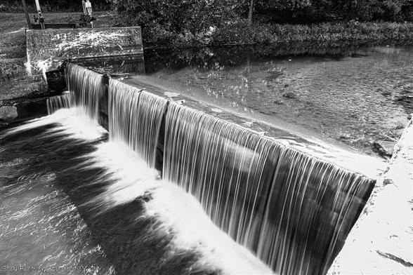 A black and white image with water flowing over an old concrete dam.