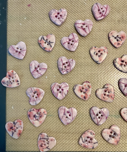A silicone mat with a scatter of two types of heart shaped buttons, one creamy pink with cherry blossoms and the other more crystalline pink with gentle marbling and veins of dark pink.