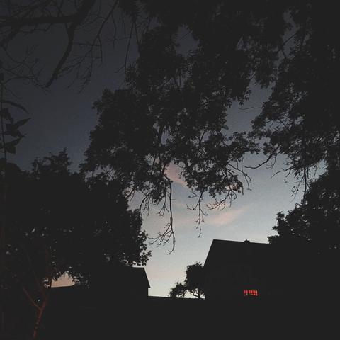 Evening sky, clouds and silhouettes of trees. A dark house with one dimly lit window.