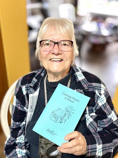 A colour photograph of an elderly woman smiling and holding a book.