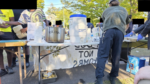 A view from behind the table. Some folks behind the table are helping serve food, while others on the other side help themselves. A crowd is visible milling around in the background.  This photo is from the Daniel's Day serve.