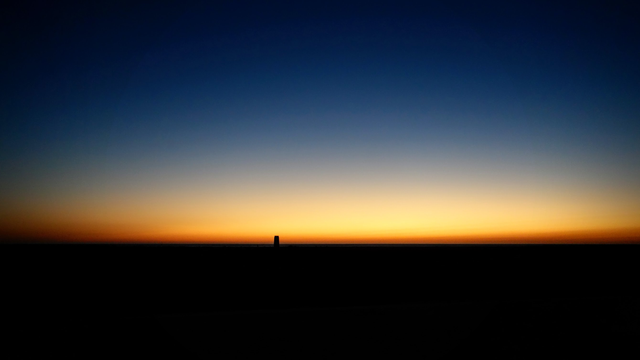Nach Sonnenuntergang, wenn es auf dem Land schon Nacht geworden ist, scheint hinter dem Horizont auf dem Meer noch der letzte Schein der Sonne. Der Himmel zeigt einen Verlauf von Nachblau über Indigo nach Blassgelb und einen schmalen Rest Sonnenorange. Hinten am Strand ist noch klein ein alter einzelner Wachturm zu erkennen.