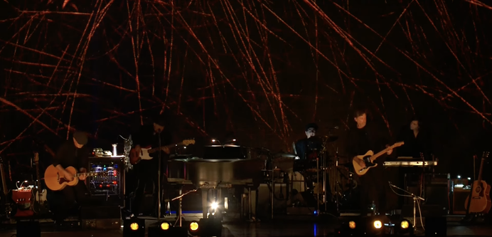 A screenshot of a rather dark stage. Two guitarists, an upright bassist, a drummer, and a keys player surround a grand piano, oriented with its keyboard facing away from the audience, Bob Dylan sitting at its keyboard behind the music stand, out of view.
