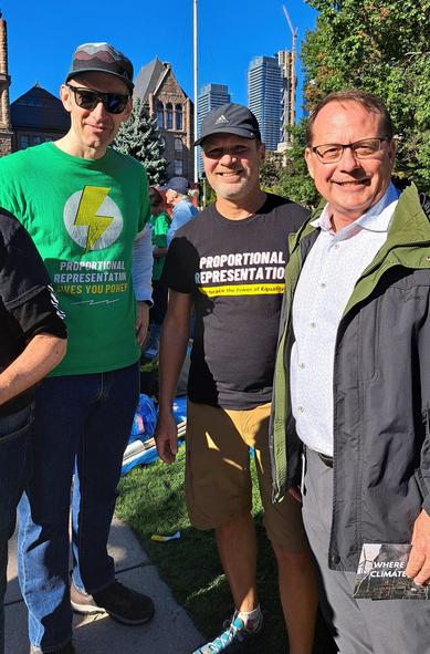 Two handsome guys in proportional representation shirts standing with Mike Schreiner. One shirt shows a lightning bolt and says proportional representation gives you power. The other guy's shirt says proportional representation, Embrace the Power of Equality.