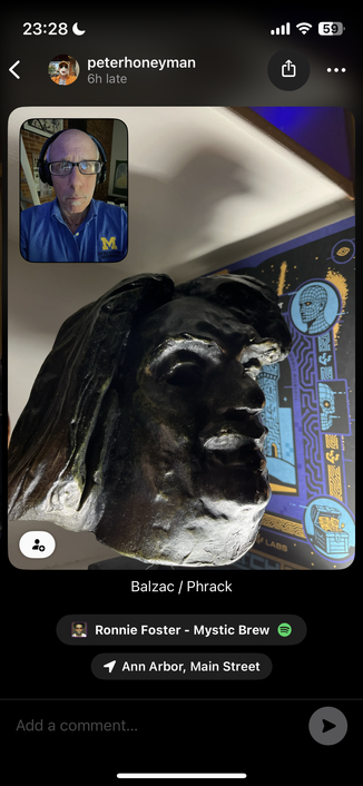 This appears to be a social media post showing what looks like a bronze or dark metal sculpture of a face in profile. The sculpture has classical features and appears to be mounted or displayed, with some colorful decorative elements visible in the background that have blue and gold coloring.

There’s a video call overlay in the top-left corner showing someone in a blue shirt (appears to be a Michigan “M” logo), and the post is labeled “Balzac / Phrack” at the bottom. The location shows “Ann Arbor, Main Street” and there’s a Spotify music indicator showing “Ronnie Foster - Mystic Brew.”

Based on the labeling and location, this appears to be a sculpture of the famous French novelist Honoré de Balzac, likely displayed somewhere on Main Street in Ann Arbor, Michigan. The post seems to be shared by someone named “peterhoneyman” and was posted “6h late” according to the timestamp.​​​​​​​​​​​​​​​​