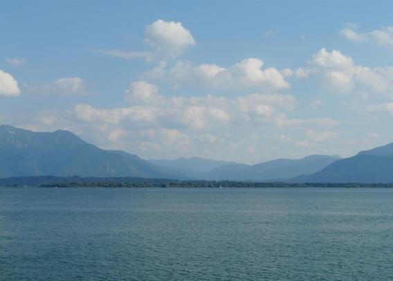 Der von Bergen umgebene Chiemsee liegt ruhig unter einem blauen Himmel mit weißen Wolken