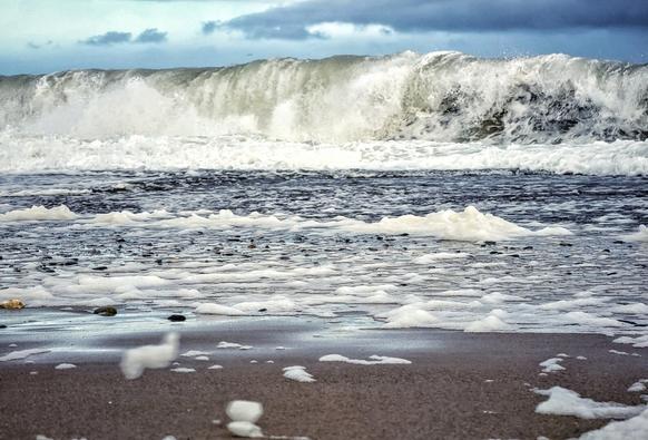 A close-up, low-angle shot of a stormy sea breaking on a sandy and rocky beach. The foreground is filled with white sea foam scattered across the wet, dark sand and smooth pebbles. In the middle ground, the receding water is a churning, foamy grey. The background is dominated by a huge, powerful wave with a white crest, crashing down just before it reaches the shore, with a moody, cloudy sky above.