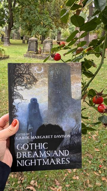 Holding the essay collection "Gothic Dreams and Nightmares" at the Bartholomew Cemetery in Göttingen. The foreground shows the book and a rosehip bush, while the background shows several old graves with sepulchral stone monuments