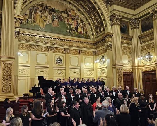 The choirs standing with folders in hands on a two level stage. The room is covered in friezes and the audience in front is clapping.
