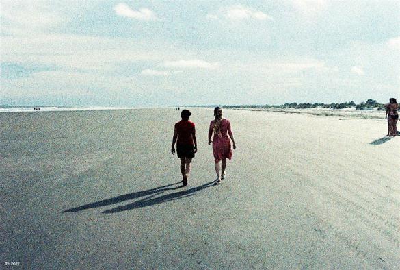 Farbfoto in den matten Farben von Adox Color Implosion. Am breiten und langen Sandstrand auf Kiawah Island sind in Bildmitte zwei Frauen mit Schatten zu sehen. Am rechten Bildrand zwei weitere Personen. Weit entfernt links ein bisschen Meer und rechts ein bisschen Dünen und Vegetation. Bassblauer Himmel mit ein paar weißen Wolken darüber. Leica R3 mit 50 mm Summilux.