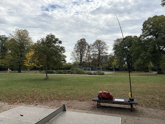 A meadow in a park with a bench and a large backpack in front, a mobile antenna mast attached to it and a soccer playground in the back