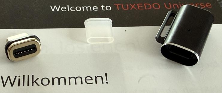 Das Bild zeigt einen USB-Typ-C-Anschluss und eine Schutzhülle, die auf einer gedruckten Karte mit der Aufschrift "Willkommen im TUXEDO-Universum" zusammen mit dem deutschen Wort "Willkommen!"