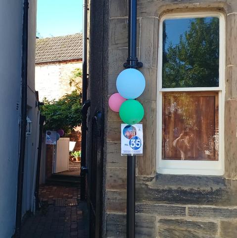 The corned of a stone house showing a window and a passage between it and the next house. Pink blue and green balloons are tied to the drainpipe with a sign reading 35