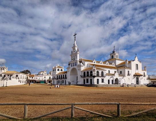A wide-angle shot of the El Rocío Sanctuary in Huelva, Spain, under a partly cloudy sky. The whitewashed church with its prominent bell tower and ornate details stands at the centre-right, facing a vast, empty dirt esplanade. A rustic wooden fence runs across the foreground, separating the viewer from the dusty ground. To the left, more white buildings extend, showing typical Andalusian architecture with balconies and terracotta roofs. Scattered figures can be seen walking around the church and esplanade.