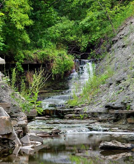 A small waterfall flows down into a pool of water in a rocky gorge in a lush green forest in Checkendon Park in Toronto, Ontario.