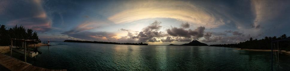photo panoramique d'un coucher de soleil sur île de Maupiti, prise depuis le motu Tiapa'a. Elle est prise depuis un ponton, à côté duquel un bateau est dans son portique, hors de l'eau. Le lagon est DZ couleur émeraude. En arrière plan, les silhouettes d'un autre motu et de l'ile principale se découpent sur le ciel éclairé des dernières lumières du jour. Des nuages de hautes altitudes reflètent encore les rayons du soleil.