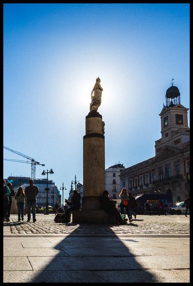 Stone monument of La Mariblanca statue on tall cylindrical column in Madrid's Puerta del Sol square, with historic clock tower building visible on right, tourists and pedestrians gathered around base, construction crane in background under clear blue sky.